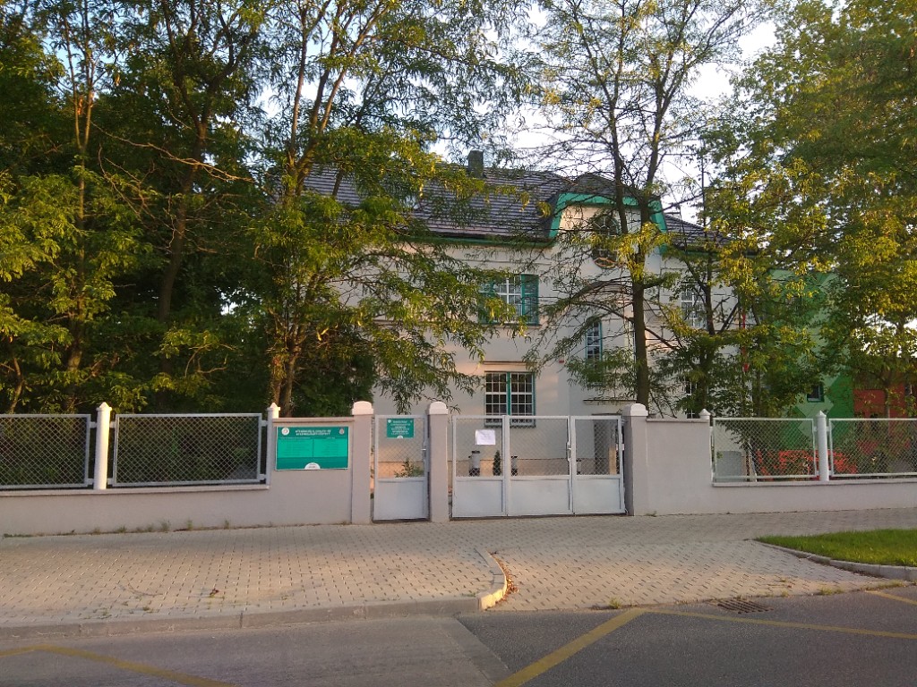 Utcai nézet kerítéssel, kapuval és a központ épületével a fák mögött